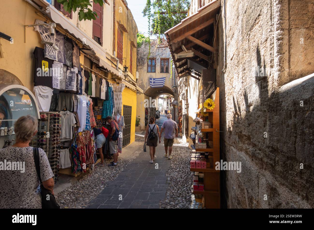 Rodi Grecia - 19 agosto 2024; turisti che camminano lungo la stretta strada della città vecchia passando per i negozi che vendono abiti alla moda tra le facciate del vecchio edificio. Foto Stock