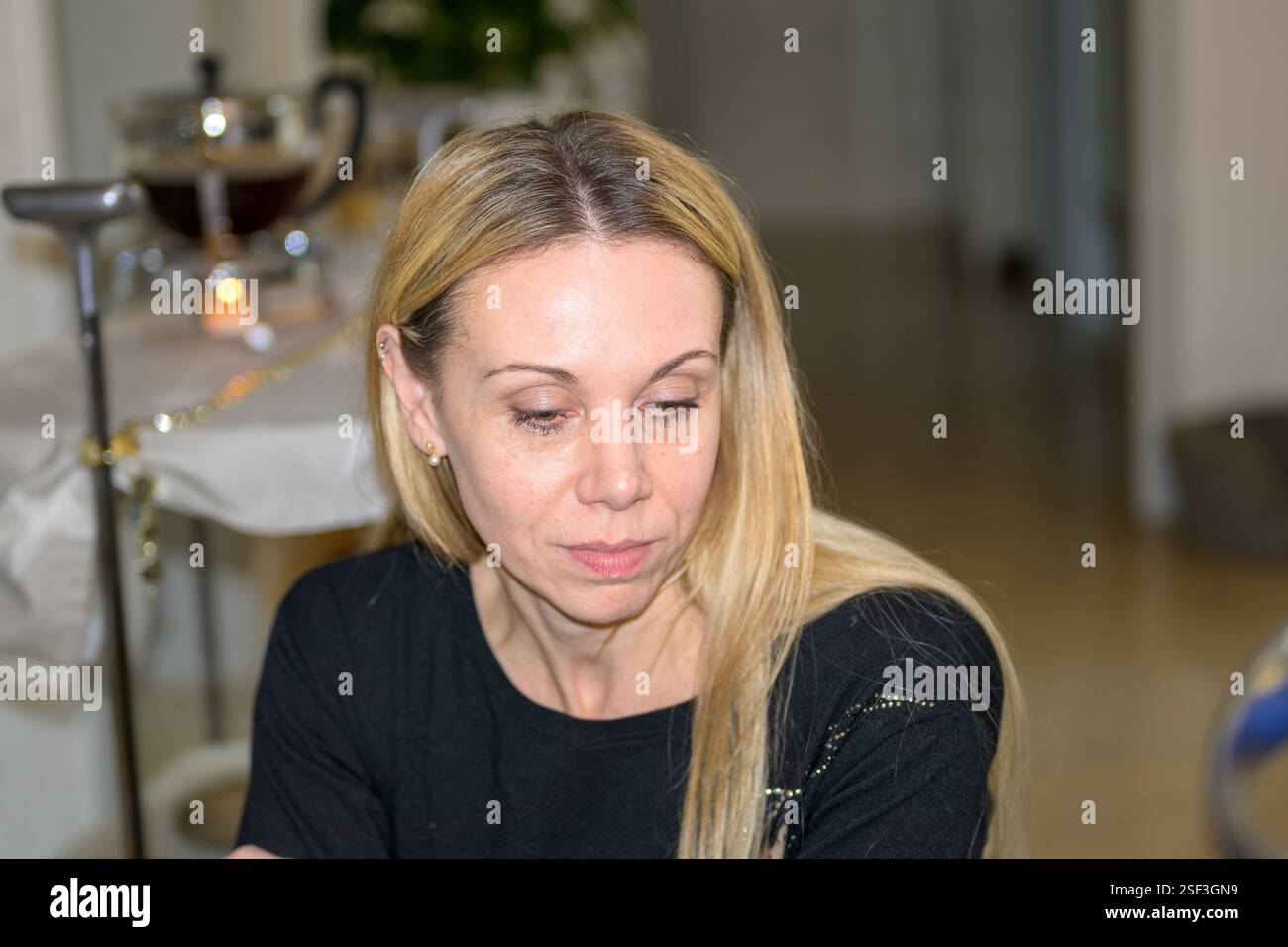 Una donna bionda dai capelli lunghi e lisci si siede in un ambiente interno e indossa una camicia nera con sottili decorazioni. Foto Stock
