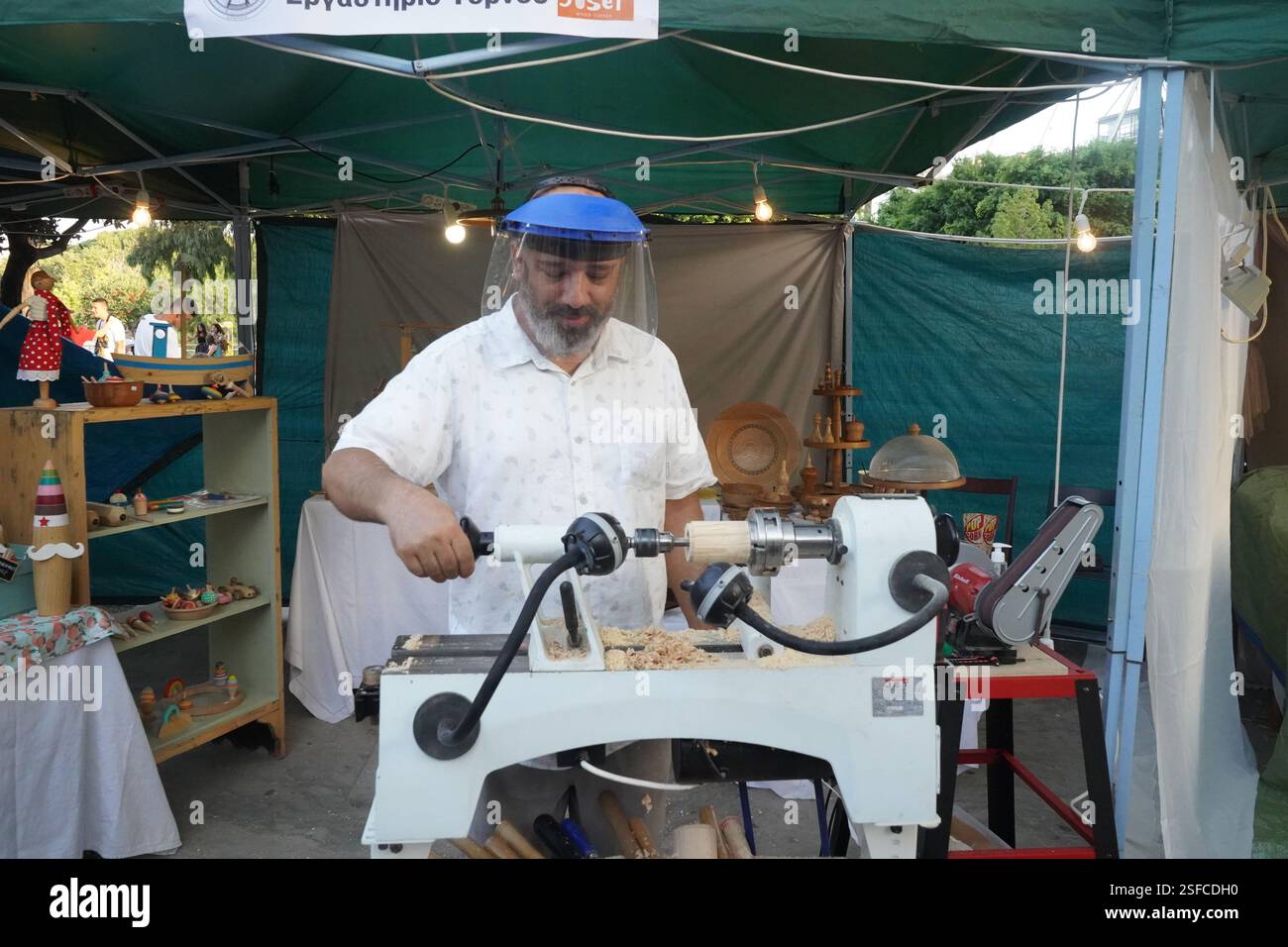 Un uomo con una visiera protettiva sta operando un tornio sotto una tenda verde. Sta girando un pezzo di legno con precisione, circondato da trucioli di legno Foto Stock