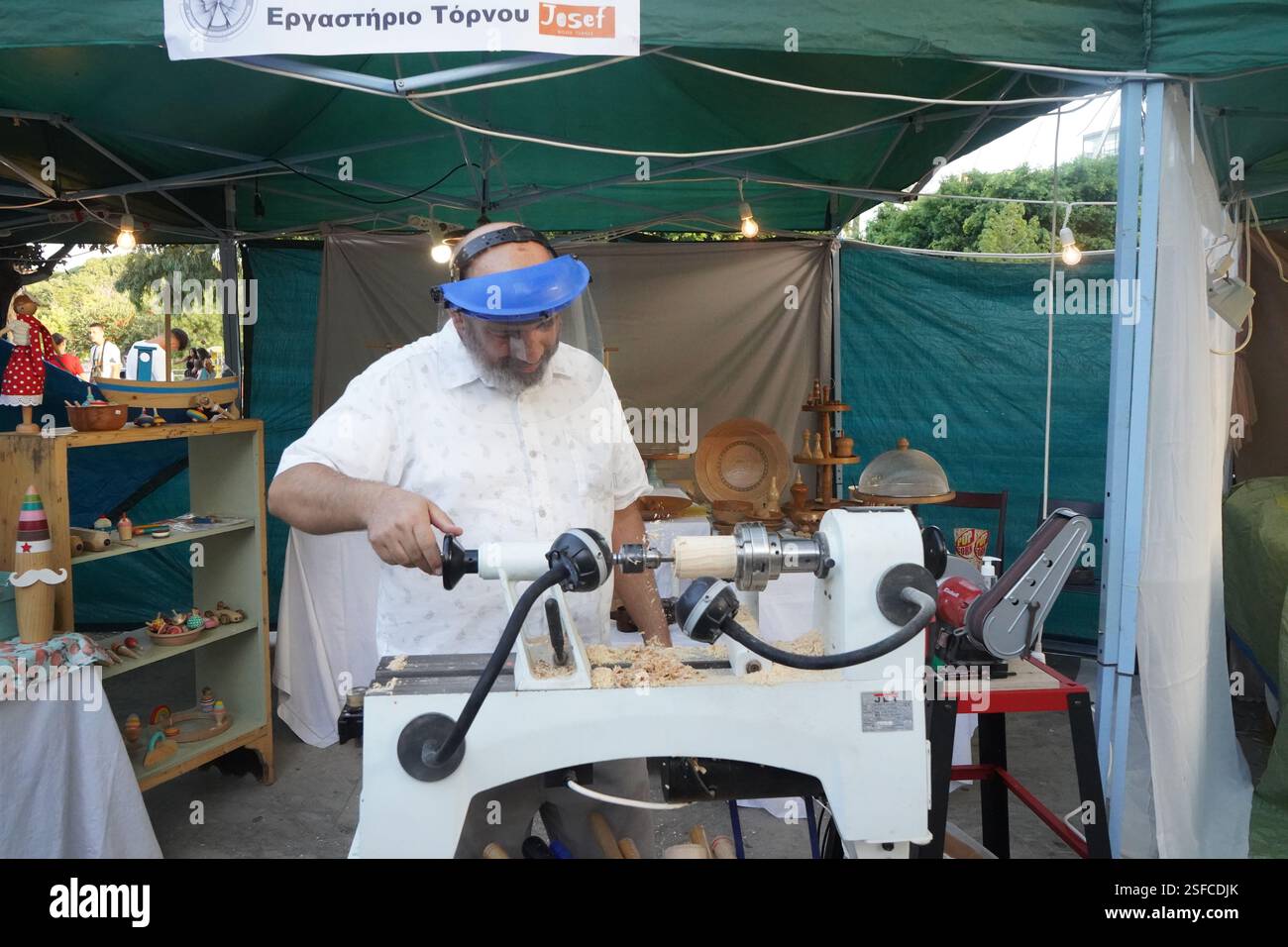 Un uomo con una visiera protettiva sta operando un tornio sotto una tenda verde. Sta girando un pezzo di legno con precisione, circondato da trucioli di legno Foto Stock