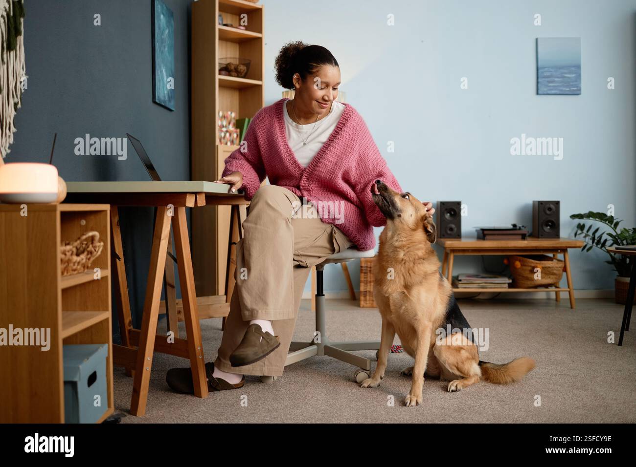 Donna seduta alla scrivania in un accogliente ufficio domestico, sorridente di un cane attento con una libreria nelle vicinanze. Camera decorata con piante e candele, a creare un'atmosfera invitante Foto Stock