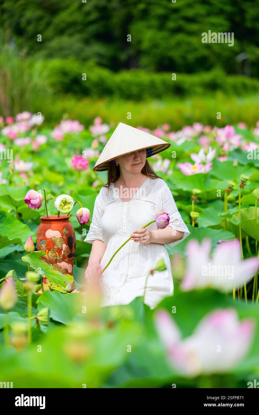 Una donna che indossa un tradizionale cappello conico vietnamita regge un fiore di loto rosa mentre è in piedi in un vivace campo di fiori di loto e foglie verdi Foto Stock