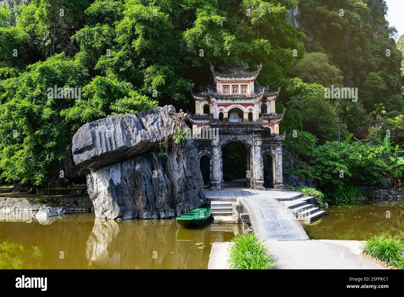 Una tranquilla vista dell'iconico ingresso della Pagoda di Bich Dong a Tam Coc, Vietnam, circondato da vegetazione lussureggiante, un laghetto riflettente e un tradizionale ponte di pietra Foto Stock