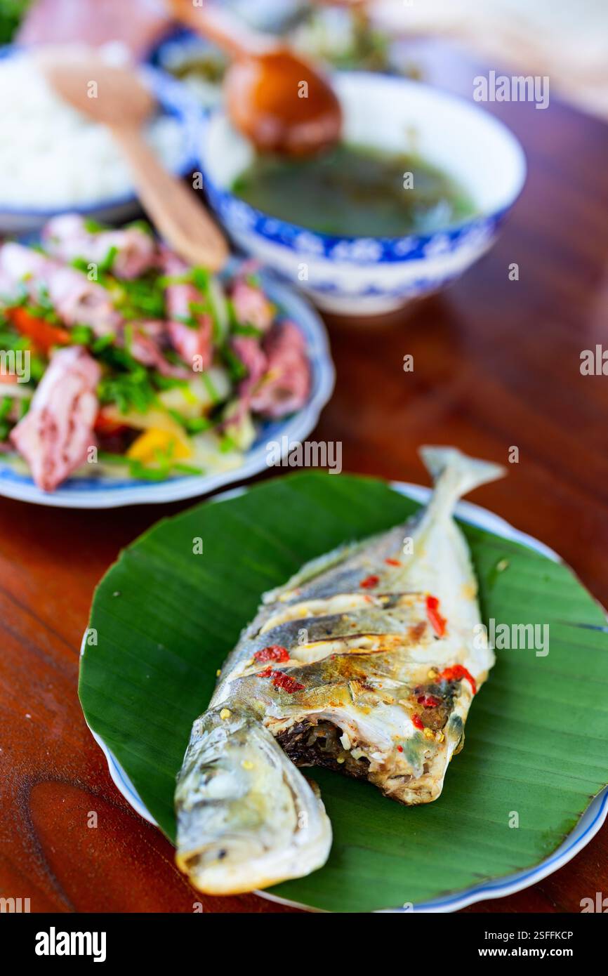 Un pesce intero alla griglia servito su una foglia di banana, accompagnato da una colorata varietà di piatti tradizionali vietnamiti, tra cui insalata fresca e zuppa Foto Stock