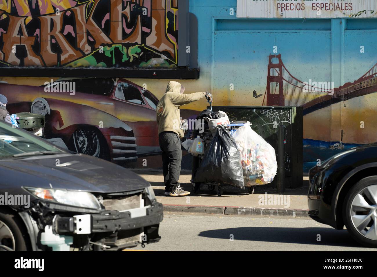 La persona raccoglie gli oggetti scartati in un carrello vicino a un murale del Golden Gate Bridge di San Francisco. Circostanze difficili. Foto Stock