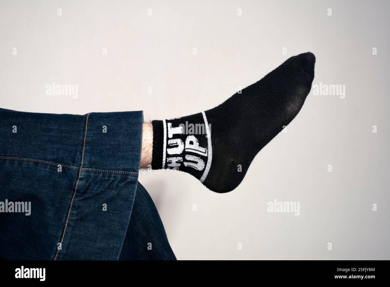 Un'immagine minimalista ma divertente con una gamba in denim, che mette in risalto un calzino nero con l'audace dichiarazione ＇Shut Up＇ in tipografia bianca. Foto Stock