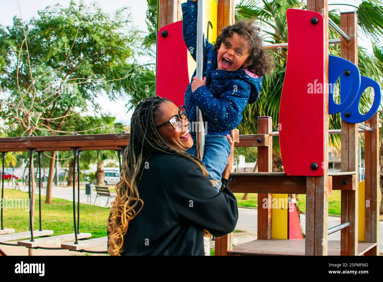 Madre che sostiene sua figlia mentre gioca su uno scivolo per parco giochi in un parco urbano, godendo del tempo di qualità insieme Foto Stock