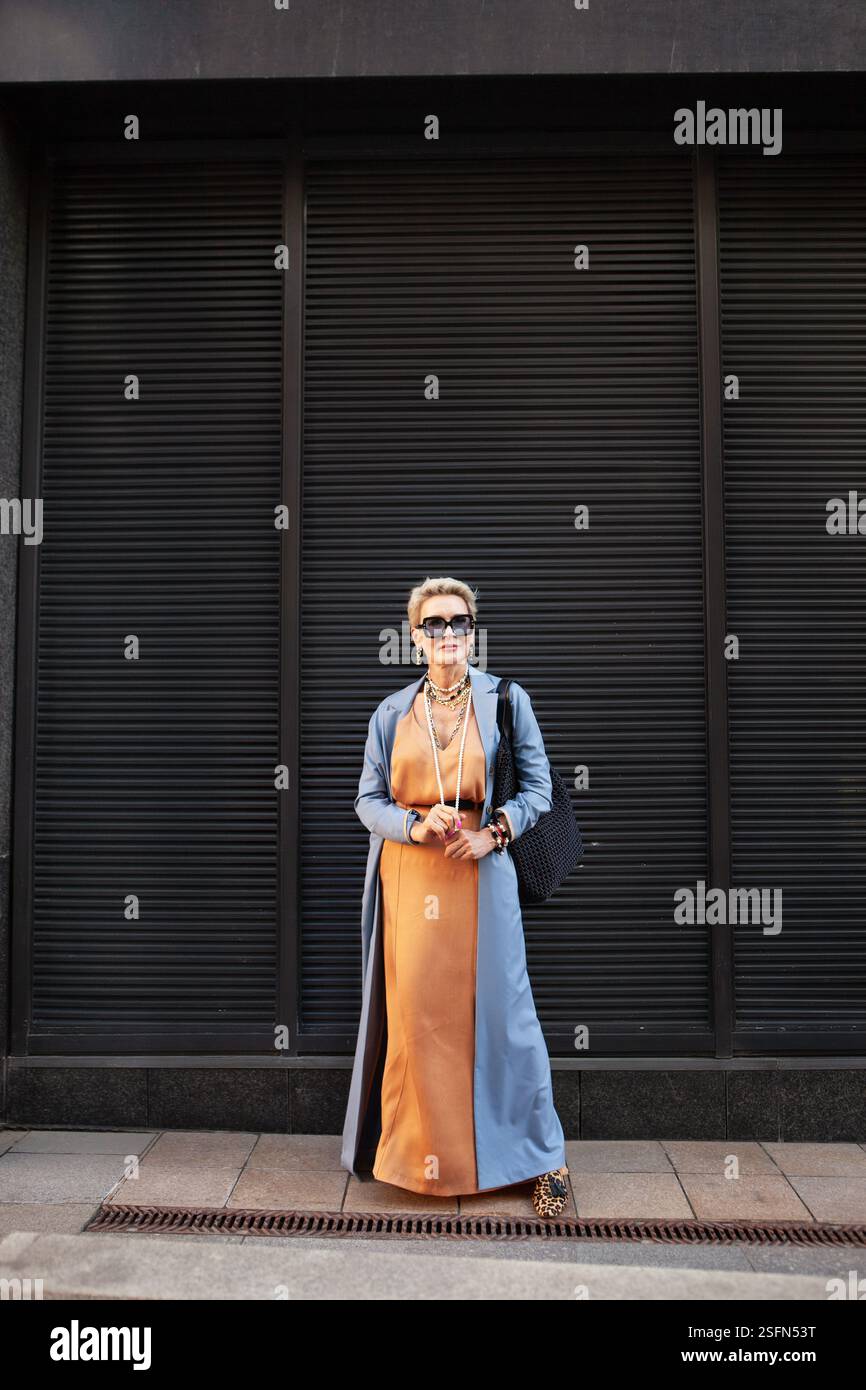 Elegante donna matura in piedi City Street con abiti alla moda, abito lungo in seta beige, cappotto blu. Foto Stock