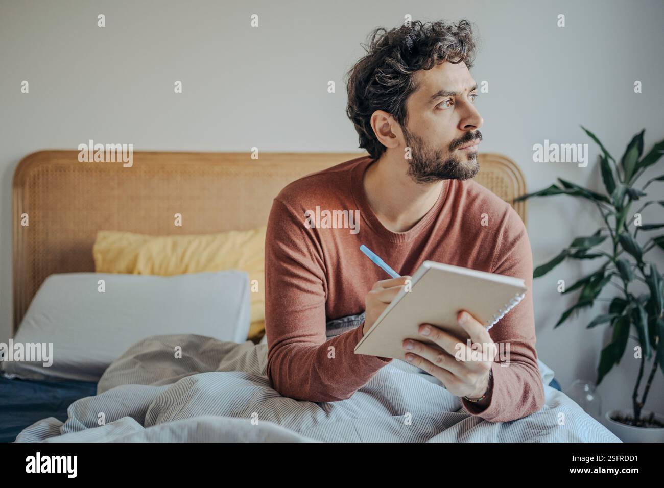 Giovane bell'uomo medio-orientale seduto a letto e a scrivere sul suo quaderno. Creatività mattutina e riflessione. Foto Stock