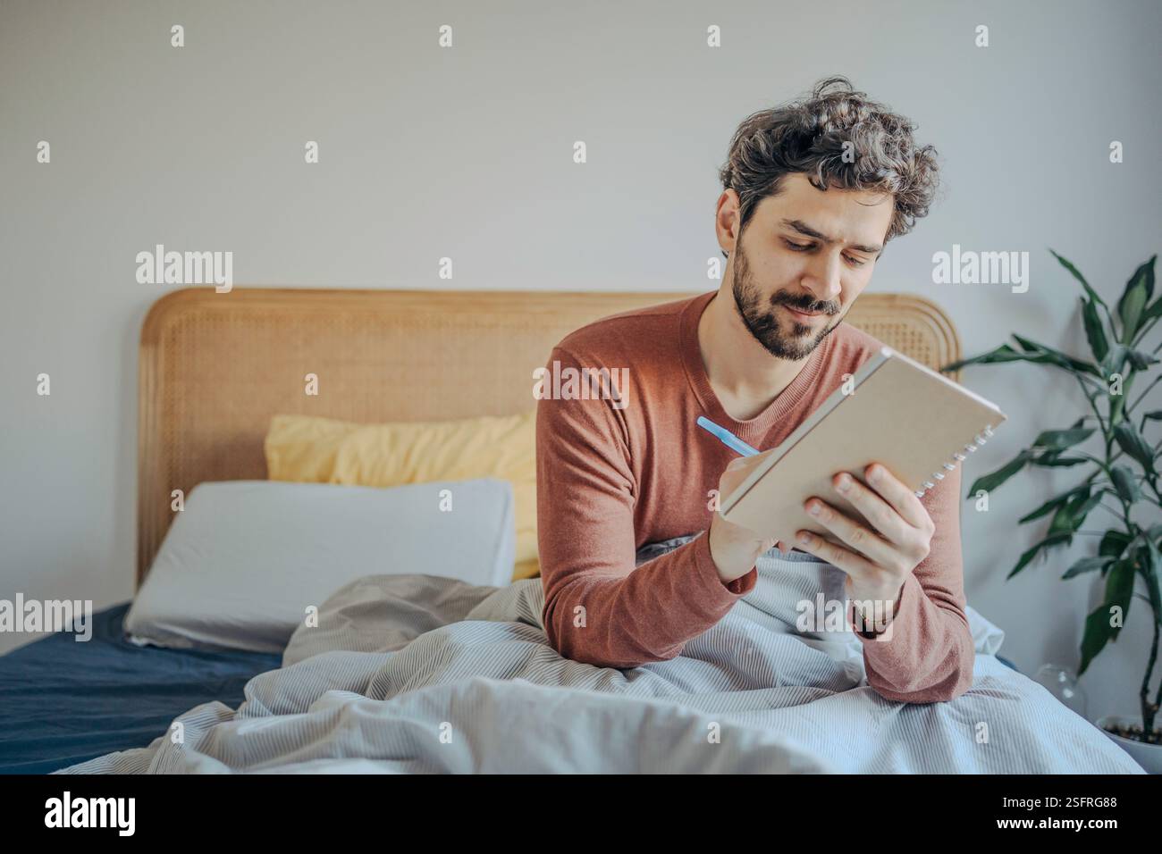 Giovane bell'uomo medio-orientale seduto a letto e a scrivere sul suo quaderno. Creatività mattutina e riflessione. Foto Stock