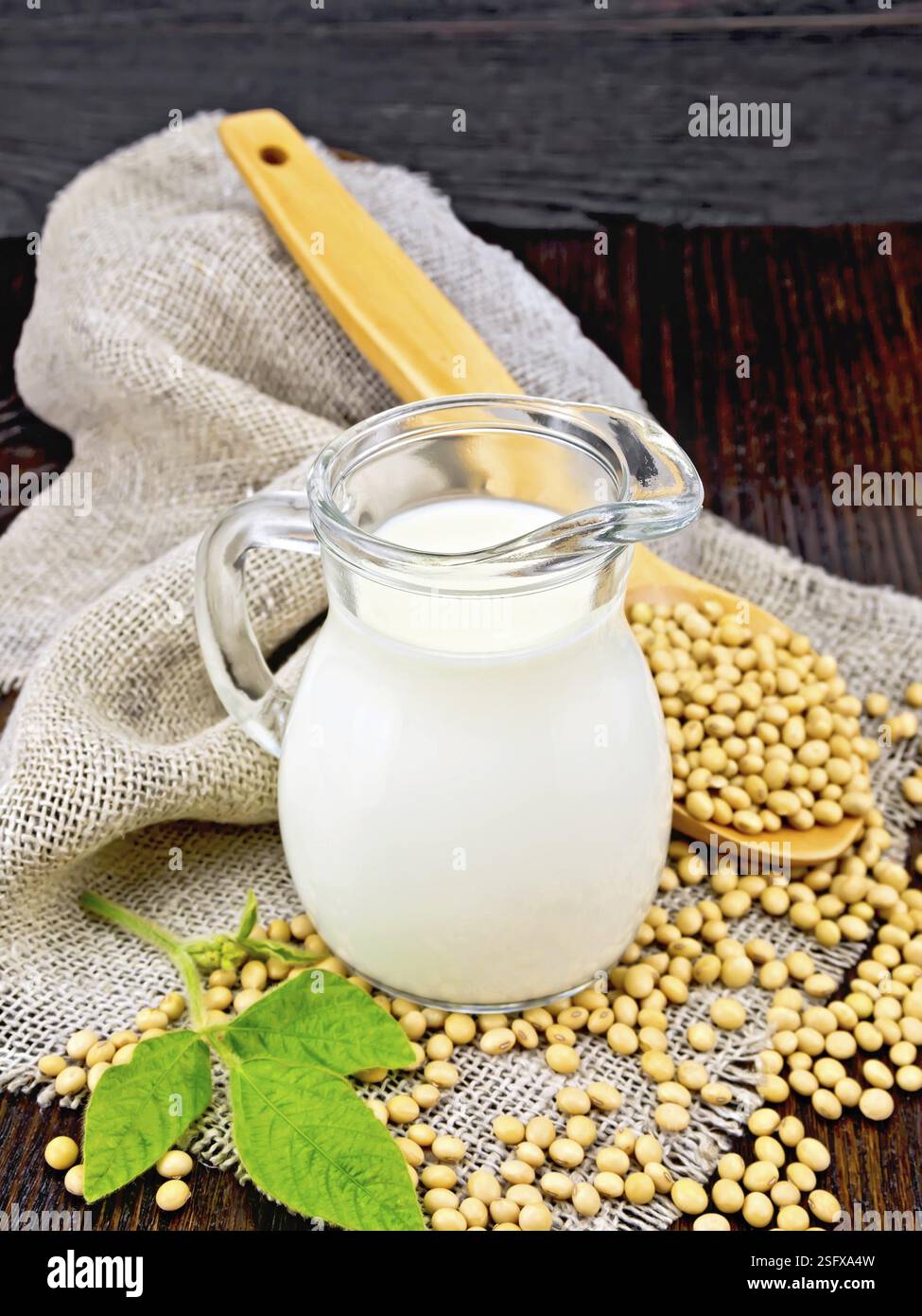 Il latte di soia in una brocca, foglia verde, i fagioli di soia in un cucchiaio su un tovagliolo di tela ruvida sullo sfondo di un oscuro pannello di legno Foto Stock
