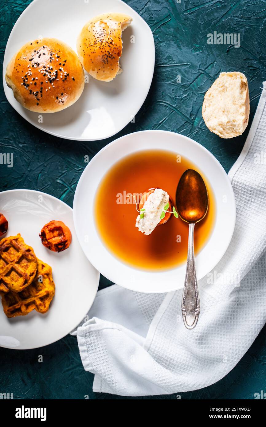 Zuppa o consomme, brodo di carne denso con carote, gnocchi di pollo serviti in un piatto bianco su sfondo verde scuro con panini di sesamo, tovagliolo Foto Stock