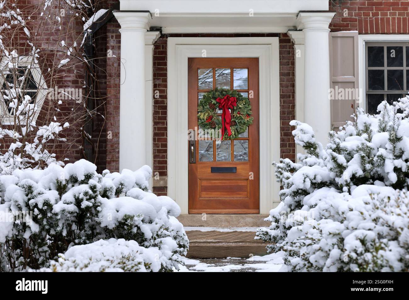 Casa tradizionale di periferia con arbusti ricoperti di neve e ghirlanda decorativa sulla porta d'ingresso Foto Stock