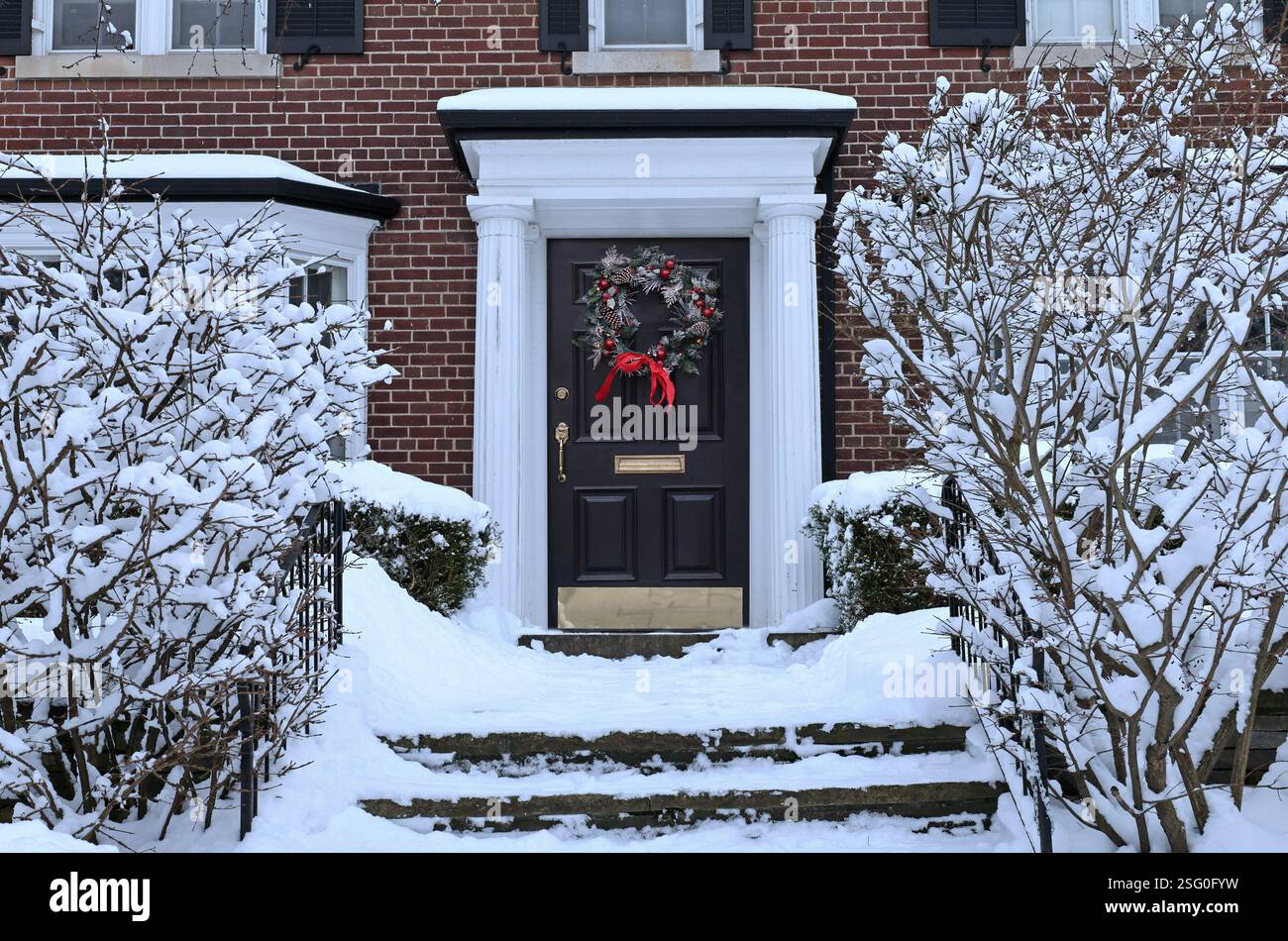 Casa tradizionale di periferia con arbusti ricoperti di neve e ghirlanda decorativa sulla porta d'ingresso Foto Stock
