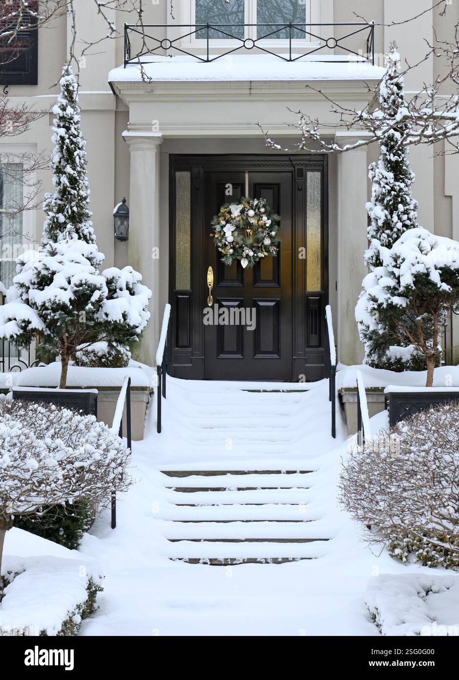 Casa tradizionale di periferia con arbusti ricoperti di neve e ghirlanda decorativa sulla porta d'ingresso Foto Stock