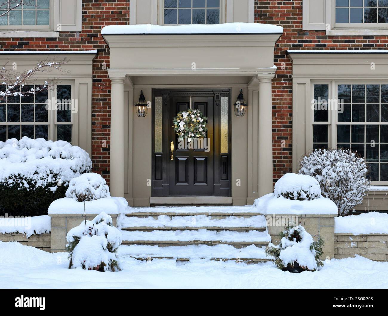 Casa tradizionale di periferia con arbusti ricoperti di neve e ghirlanda decorativa sulla porta d'ingresso Foto Stock