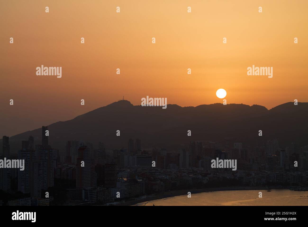 Un tramonto sereno sullo skyline della città con montagne sullo sfondo. Il sole e' grande e arancione, che proietta un caldo bagliore sugli edifici e sull'acqua. Foto Stock