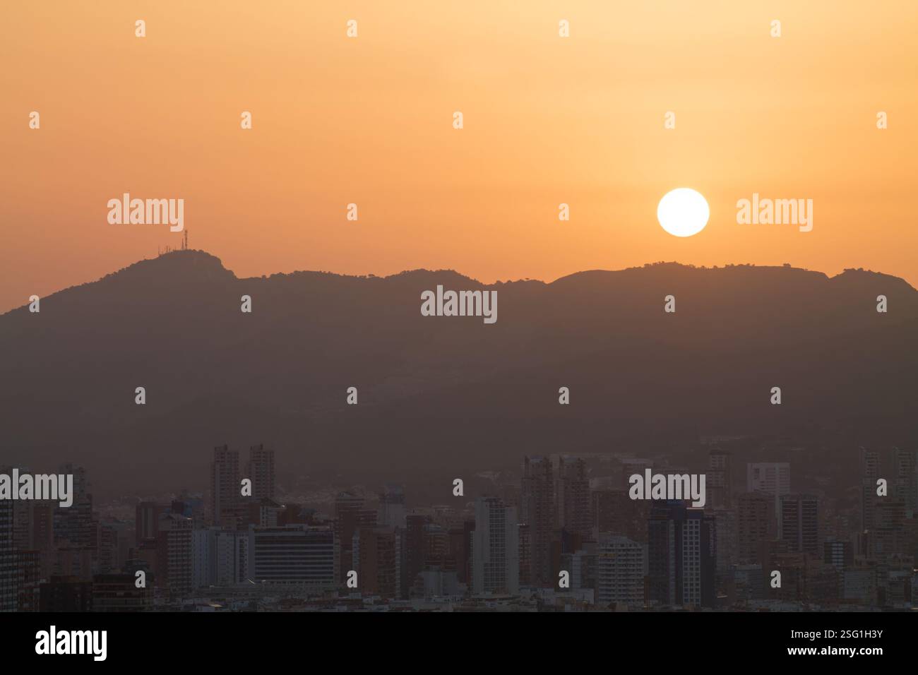 Un tramonto sereno sullo skyline della città con montagne sullo sfondo. Il sole e' grande e arancione, che proietta un caldo bagliore sugli edifici. L'atmosfera Foto Stock