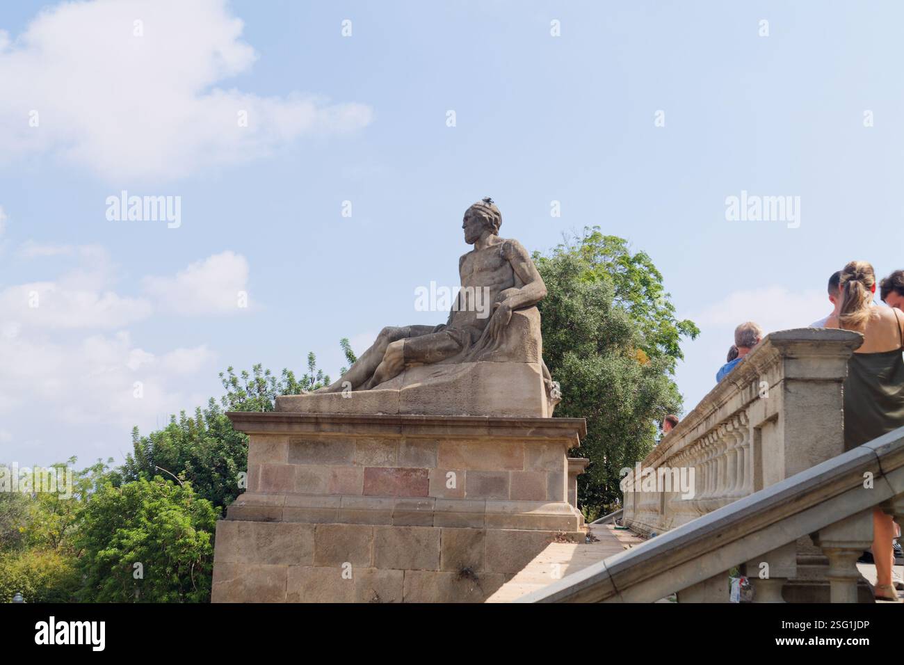 Una statua in pietra di una figura seduta, circondata da vegetazione e persone su una scala. La statua è dettagliata e sembra essere parte di un arcata più grande Foto Stock