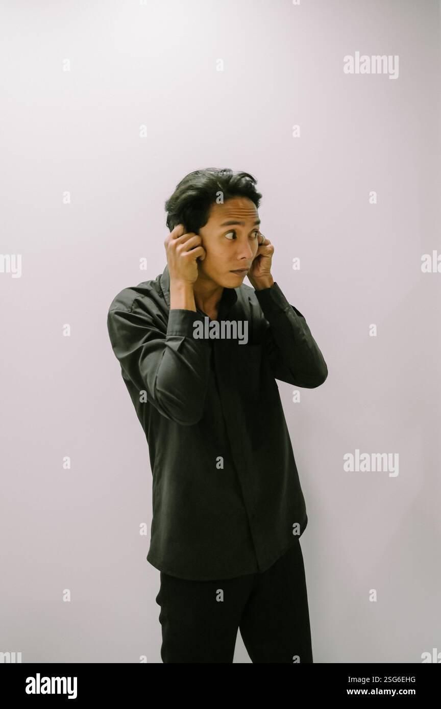 Uomo che copre le orecchie con le mani, camicia nera, sfondo semplice. Foto Stock