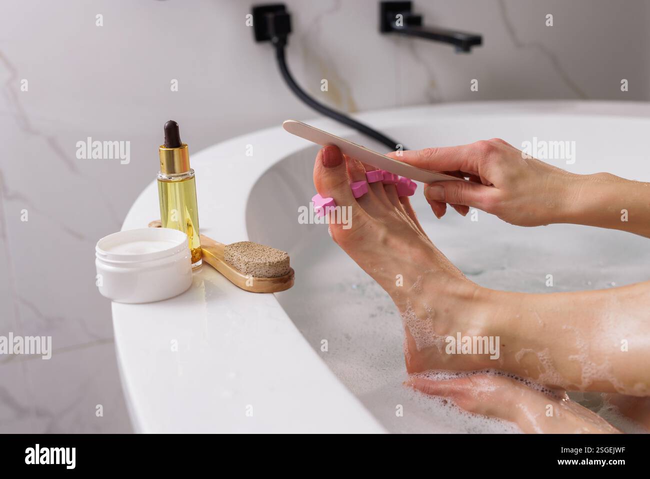Cura di sé a casa: Donna che si gode una rilassante pedicure in una vasca da bagno, che filtra le unghie dei piedi con separatori rosa. Prodotti essenziali per il centro benessere come panna, olio e pu Foto Stock