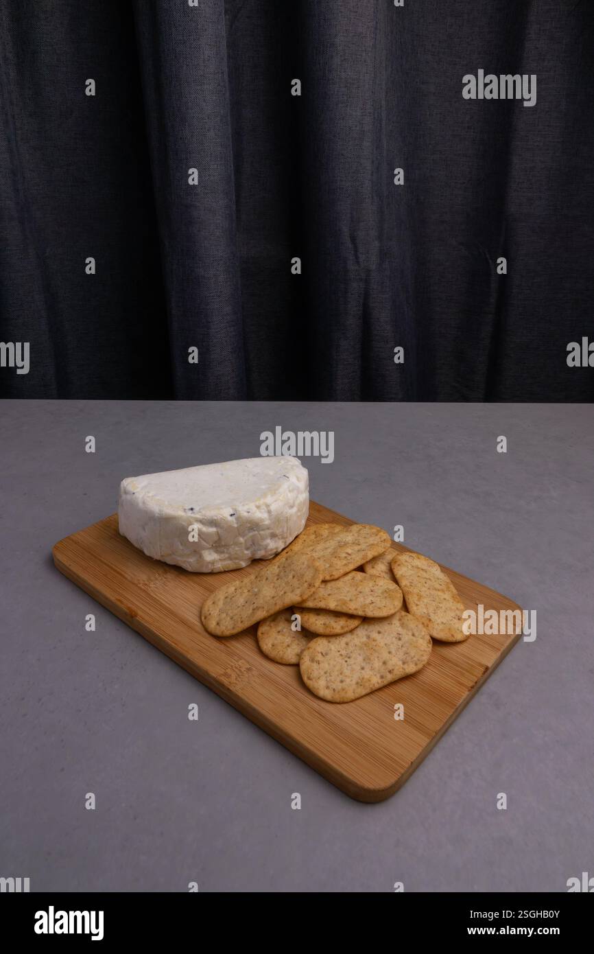 Un pezzo rotondo di formaggio siede su un tavolo da servizio di legno accanto a diversi cracker, il tutto adagiato su uno sfondo scuro. Foto Stock