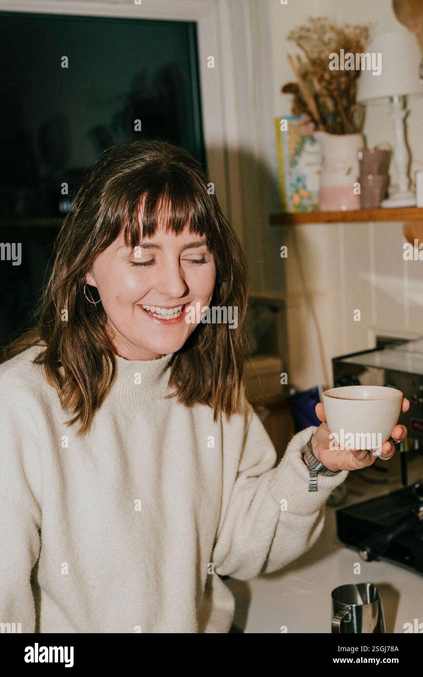 Una donna sorride mentre tiene una tazza di caffè. Indossa un maglione bianco e i capelli vengono tirati indietro. La scena si svolge in una cucina con Foto Stock
