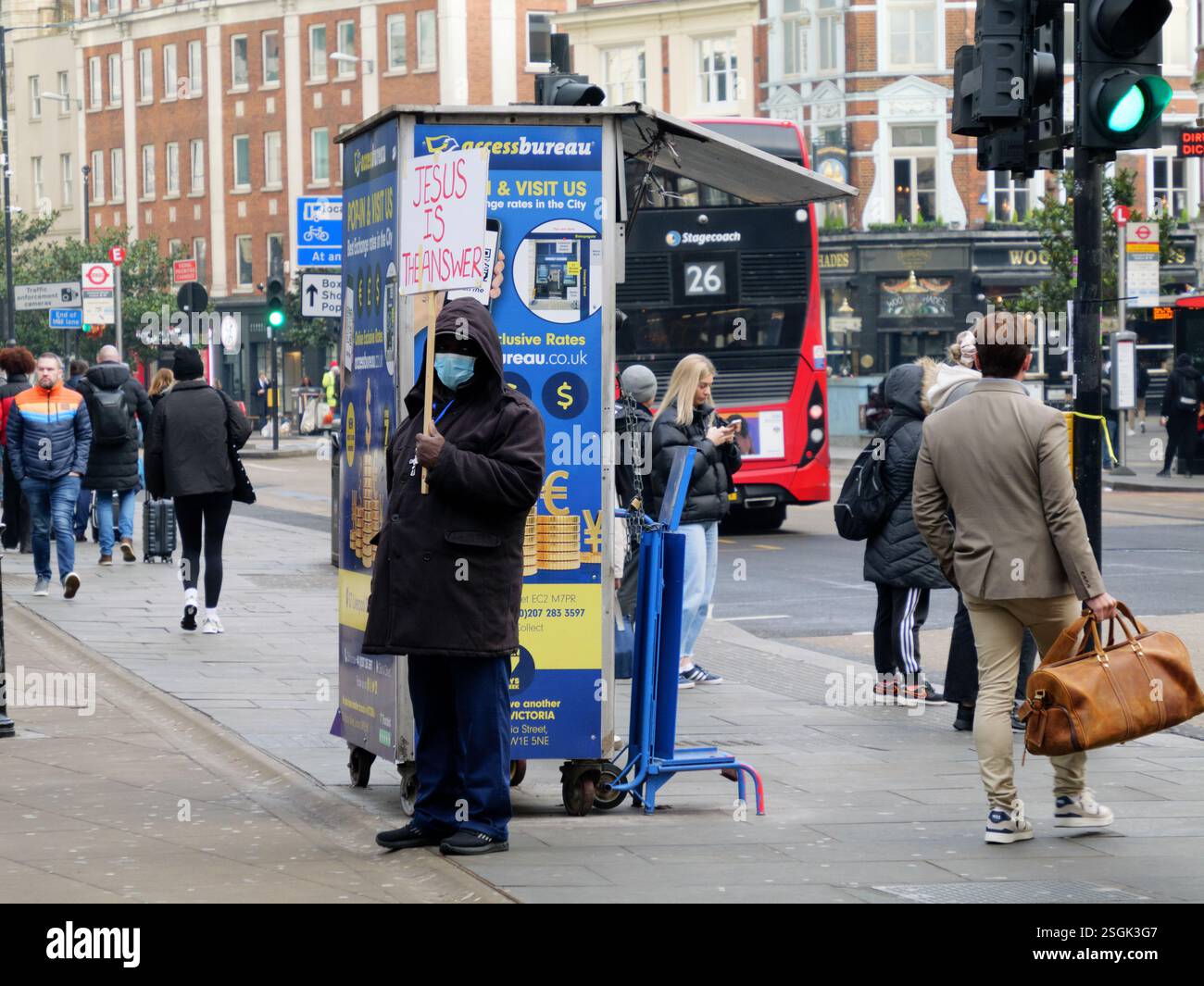 Un predicatore cristiano si trova fuori dalla stazione di Liverpool Street, Londra, Regno Unito, con un cartello che recita "Gesù è la risposta" mentre i pendolari passano davanti, immersi nella loro routine quotidiana Foto Stock