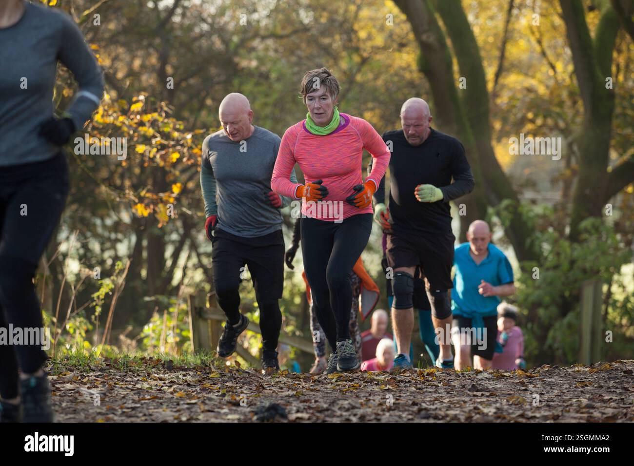 Un gruppo di persone che corrono in salita in una foresta autunnale, indossando outfit atletici dai colori vivaci. Sale Water Park, Manchester, Regno Unito Foto Stock