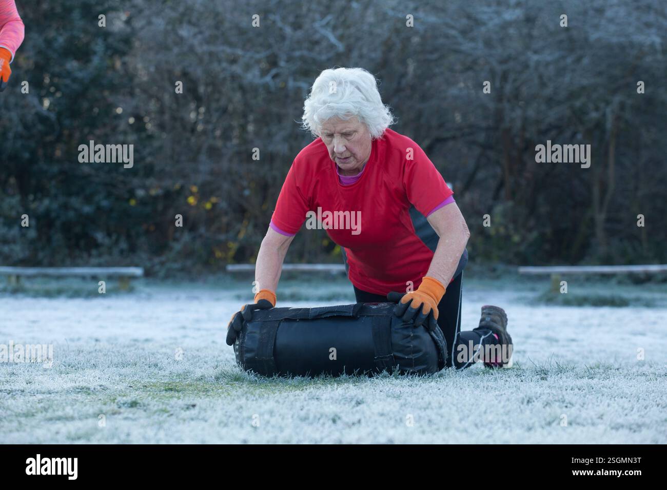 Donna anziana che si inginocchia su un campo ghiacciato, indossa una camicia rossa e guanti arancioni, solleva un sacco di sabbia nero durante un allenamento all'aperto. Sale Water Park, Manchester, Regno Unito Foto Stock