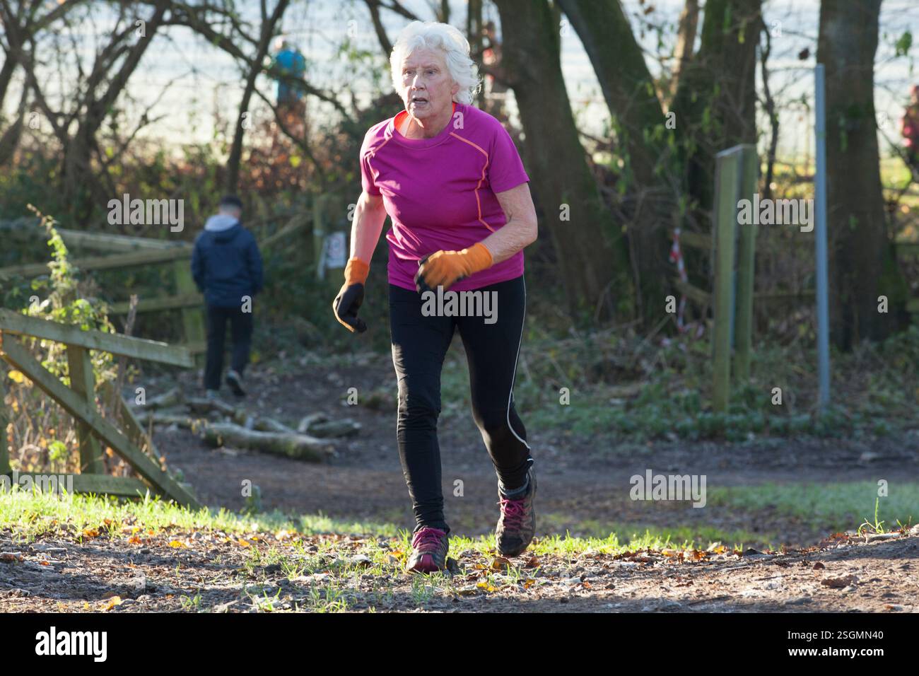Donna anziana che fa jogging in salita su un sentiero nella foresta, indossa una camicia rosa brillante, pantaloni neri e guanti, sale Water Park, Manchester, REGNO UNITO Foto Stock