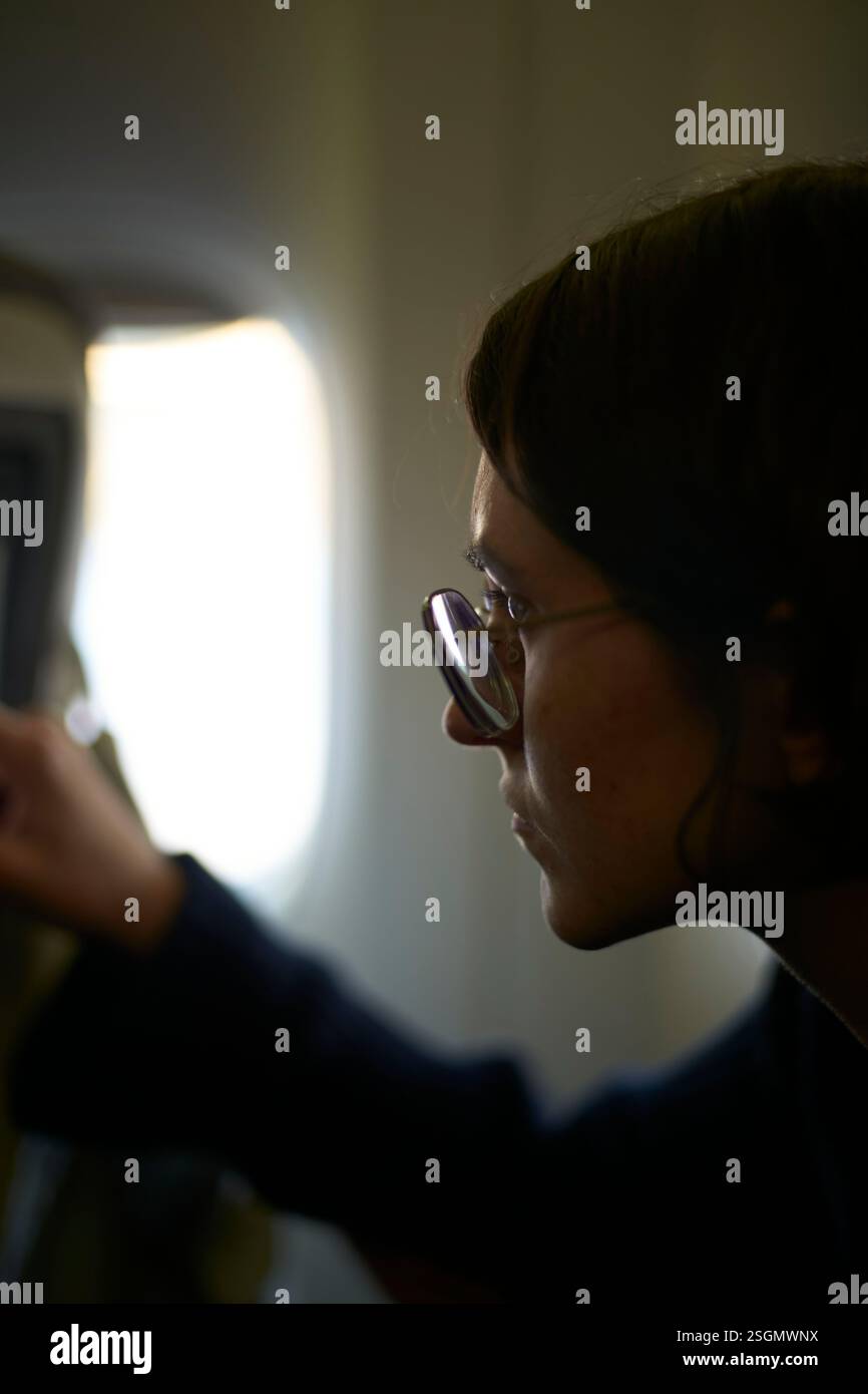 Donna che indossa gli occhiali si concentra con attenzione sullo schermo con dietro la finestra dell'aereo sfocata Foto Stock