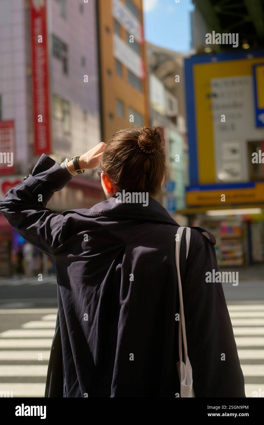 Donna con un cappotto nero e una borsa bianca attraversa una strada soleggiata circondata da edifici alti. Tokyo, Giappone Foto Stock