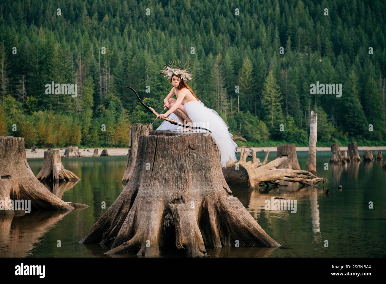 Una donna con un abito bianco, un arco, una freccia e una corona si posa su un ceppo d'albero in una scenografica cornice di un lago boscoso. Rattlesnake Lake, Puget Sound, Washington, USA Foto Stock