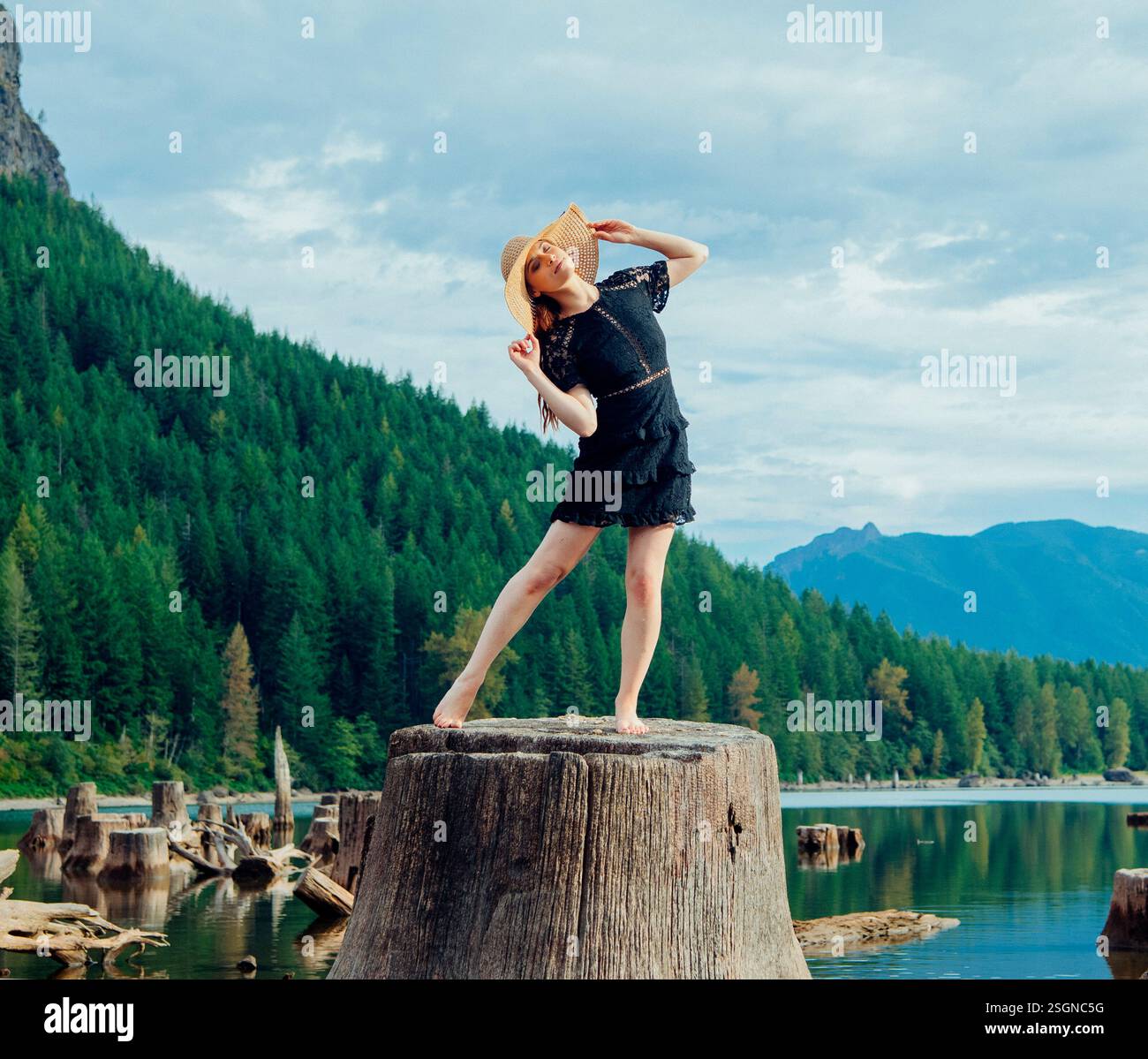 Una donna in abito nero e cappello posa su un ceppo di alberi tra montagne panoramiche e lago. Rattlesnake Lake, Puget Sound, Washington, USA Foto Stock