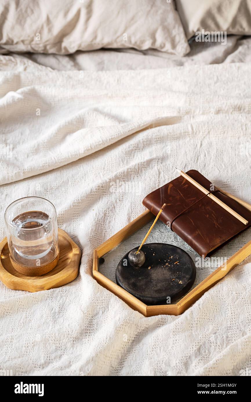 Bastoncino di incenso con fumo, bicchiere d'acqua e quaderno sul letto. Scena rituale mattutina. Foto Stock