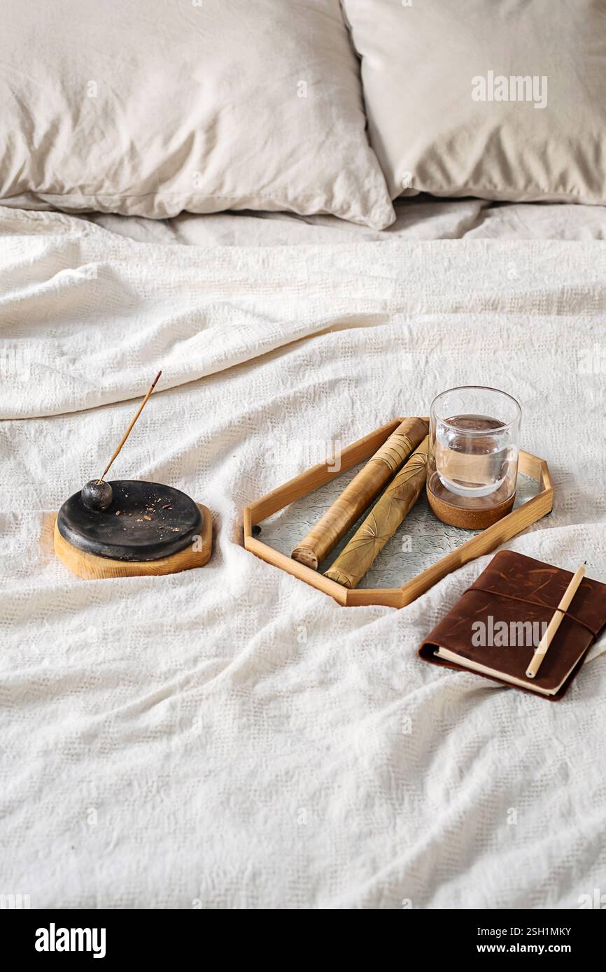 Bastoncino di incenso con fumo, bicchiere d'acqua e quaderno sul letto. Scena rituale mattutina. Foto Stock