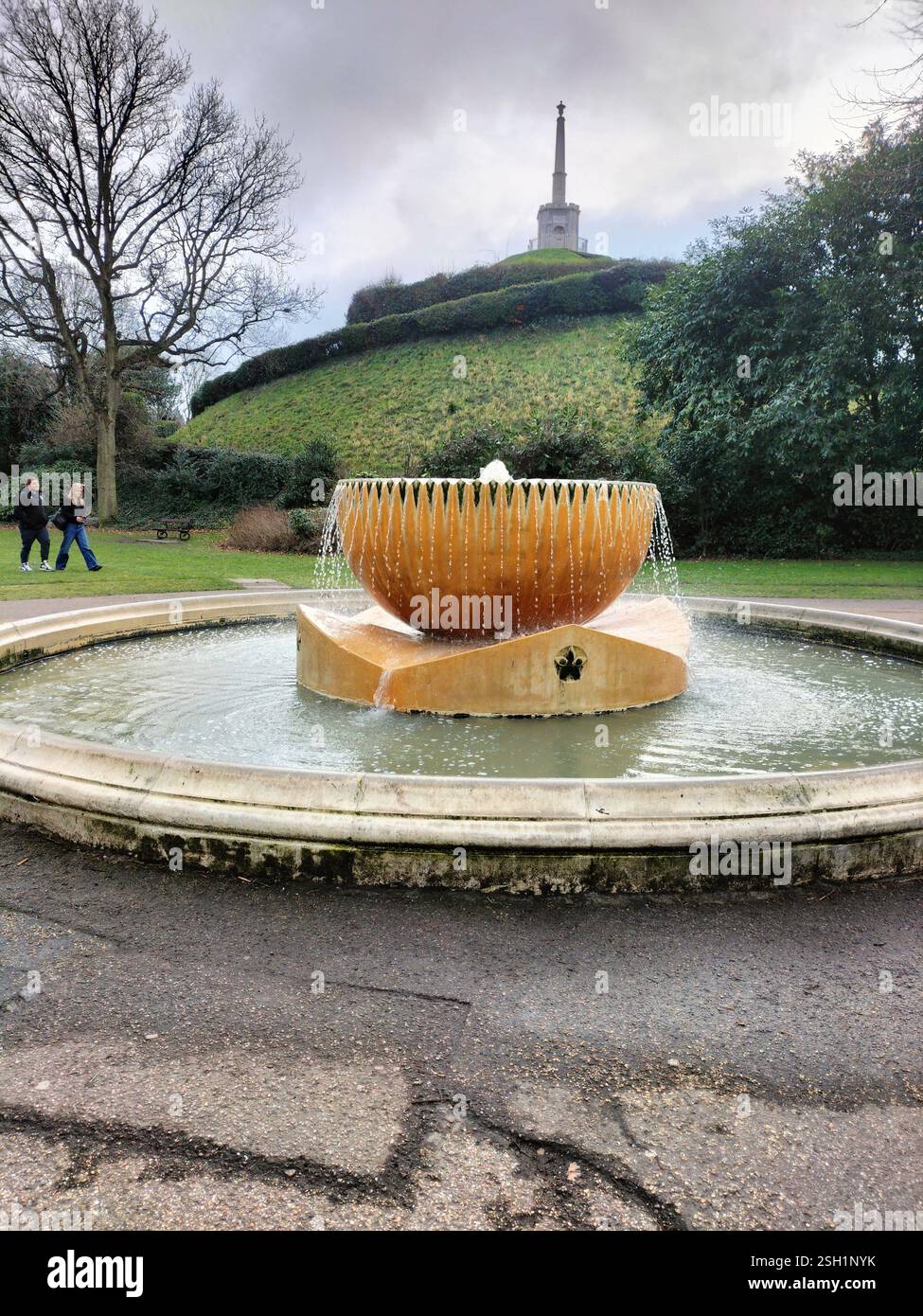 Una fontana decorativa si trova al centro di un parco, con i visitatori che passeggiano nelle vicinanze. Dietro la fontana sorge una collina erbosa. - Immagine stock catturata con smartphone