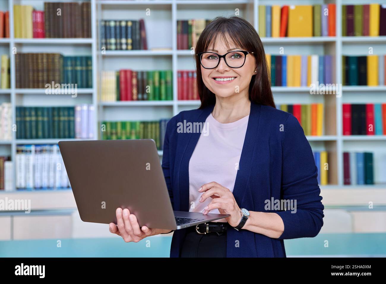 Ritratto di una donna sorridente di mezza età professoressa con laptop all'interno della biblioteca Foto Stock