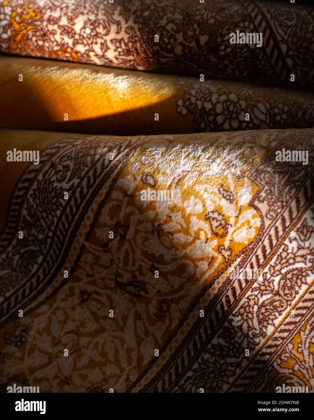 Primo piano di un tradizionale tappeto persiano con intricati motivi floreali in calde tonalità oro e marrone, illuminato dalla luce soffusa del sole Foto Stock