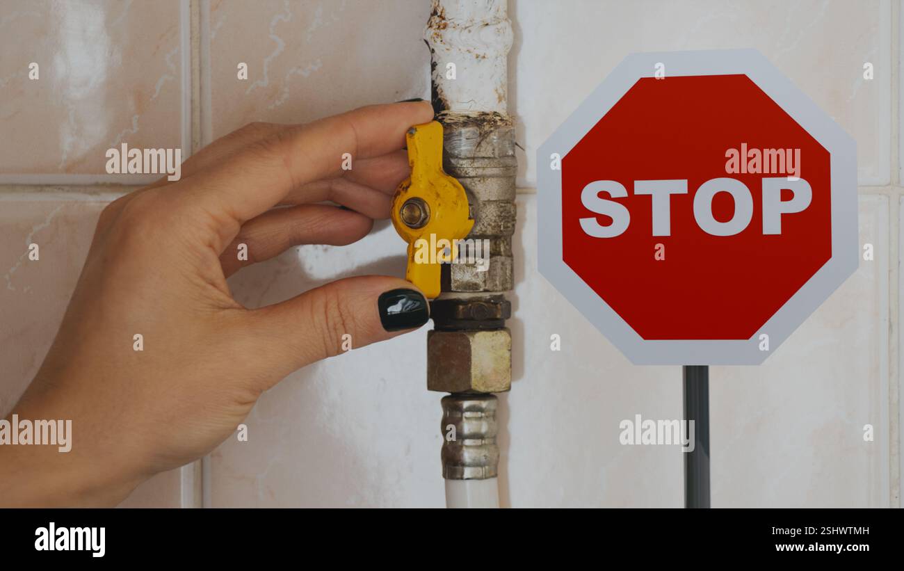 La mano di una donna chiude il rubinetto su un tubo del gas in un appartamento e accanto ad esso si trova un segnale di stop, un divieto e un rifiuto di gas e sanzioni per il gas Foto Stock