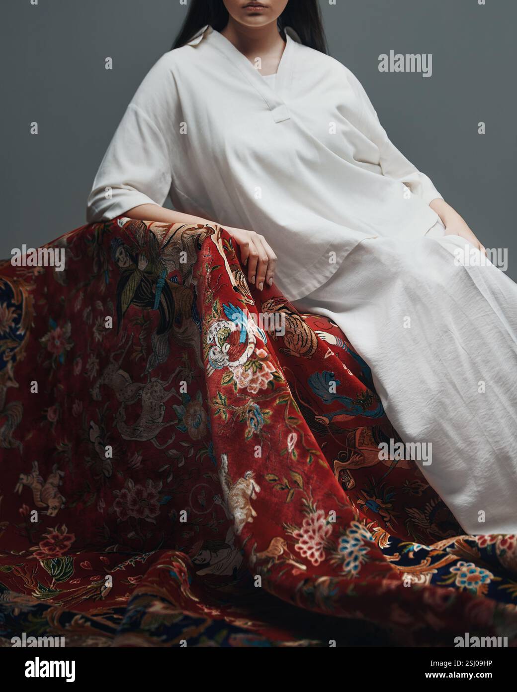 Donna in bianco drappeggiata in un tappeto rosso con creature mitiche e motivi floreali, che fondono moda e arte culturale Foto Stock