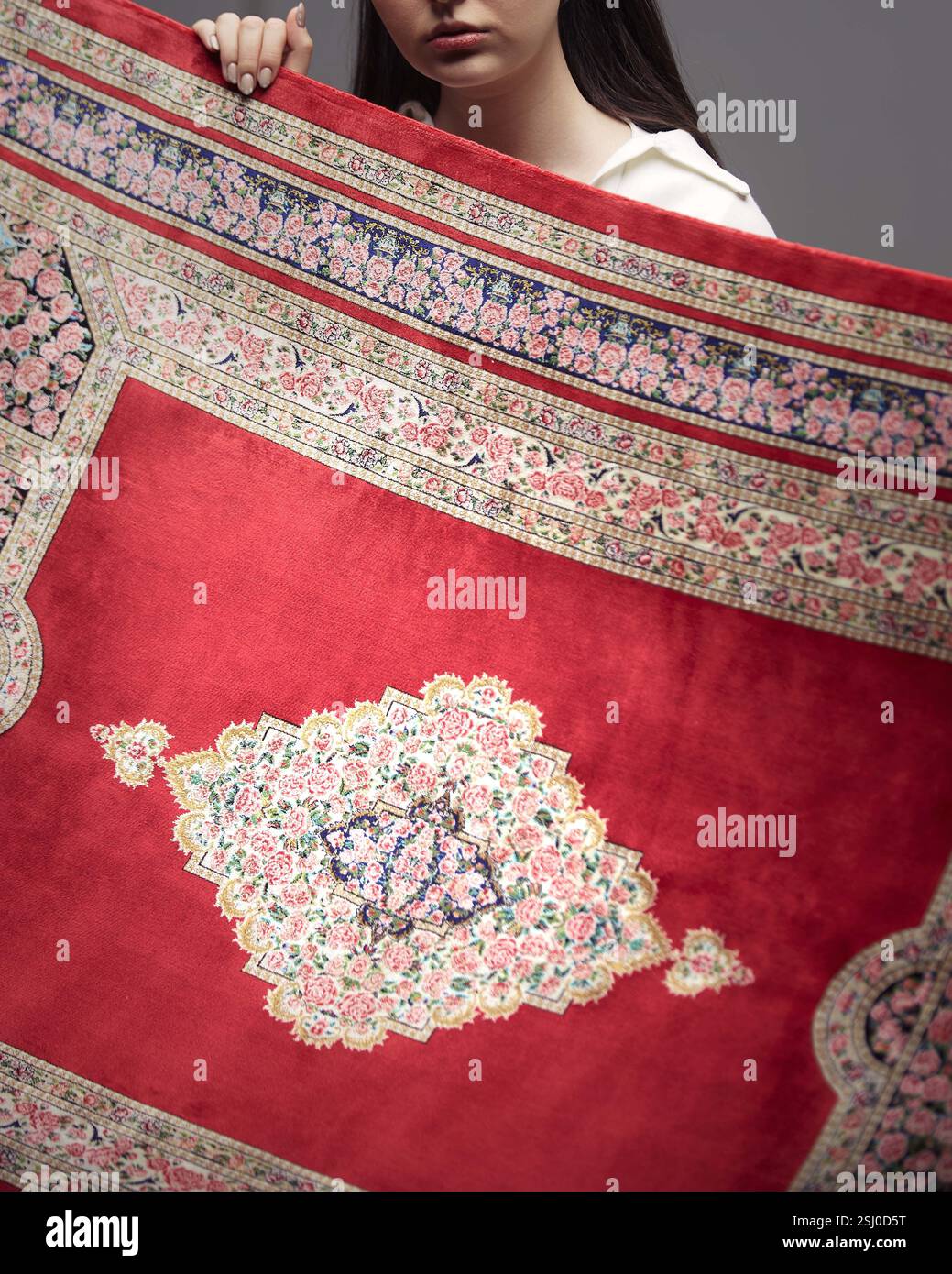 Il vivace tappeto floreale rosso si dispiega, rivelando il suo design ornato e i colori ricchi, tenuto da una donna in un ambiente studio. Foto Stock