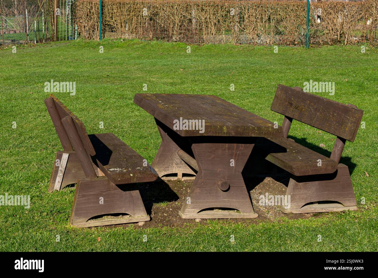 Un rustico tavolo da picnic in legno siede su un lussureggiante prato verde in un parco. Il sole splende brillantemente, proiettando ombre morbide sul terreno. Le boccole vicine forniscono un Foto Stock