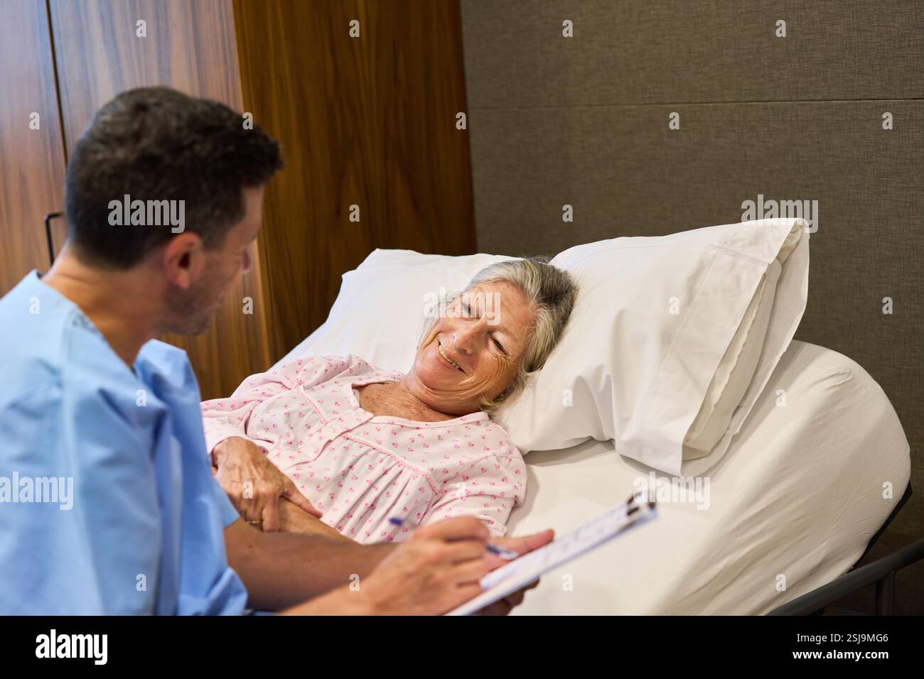 Un operatore sanitario comunica con una donna anziana che riposa in un letto di una struttura di riabilitazione, simboleggiando l'assistenza medica, la compassione e il sup del paziente Foto Stock