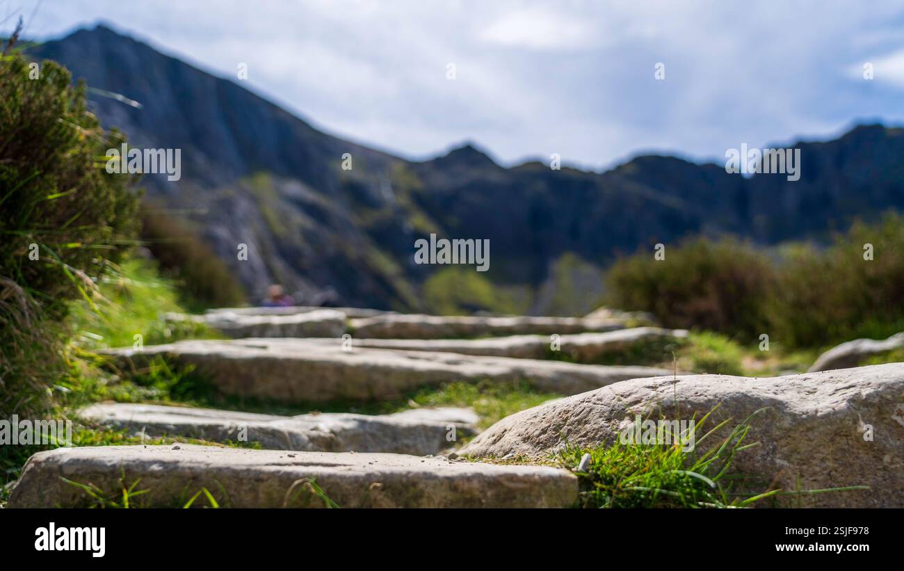 Primo piano degli antichi gradini di pietra che conducono a Llyn Idwal: Un percorso robusto incorniciato da montagne fuori fuoco e vegetazione sparsa Foto Stock