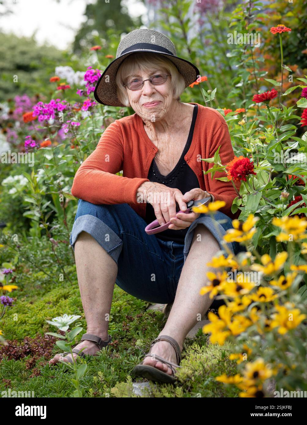 Ritratto della donna adulta più anziana giardiniera seduta nel suo vivace giardino pieno di fiori in fiore. Sorride contentemente. Foto Stock