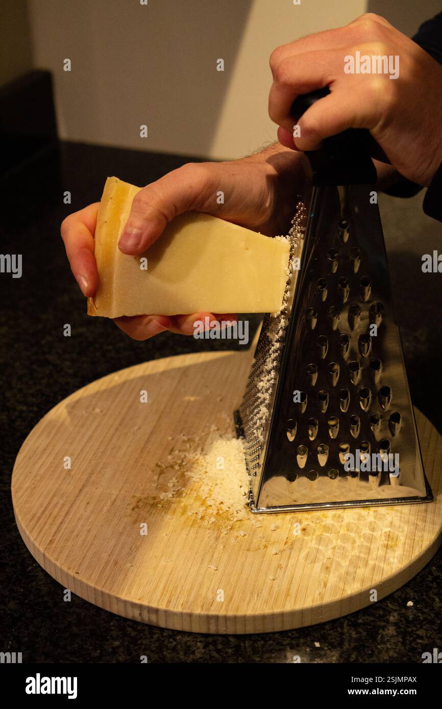 Un primo piano di una mano che utilizza una grattugia per macinare il parmigiano fresco, con il formaggio che cade su una scrivania, mostrando un processo culinario in azione. Foto Stock