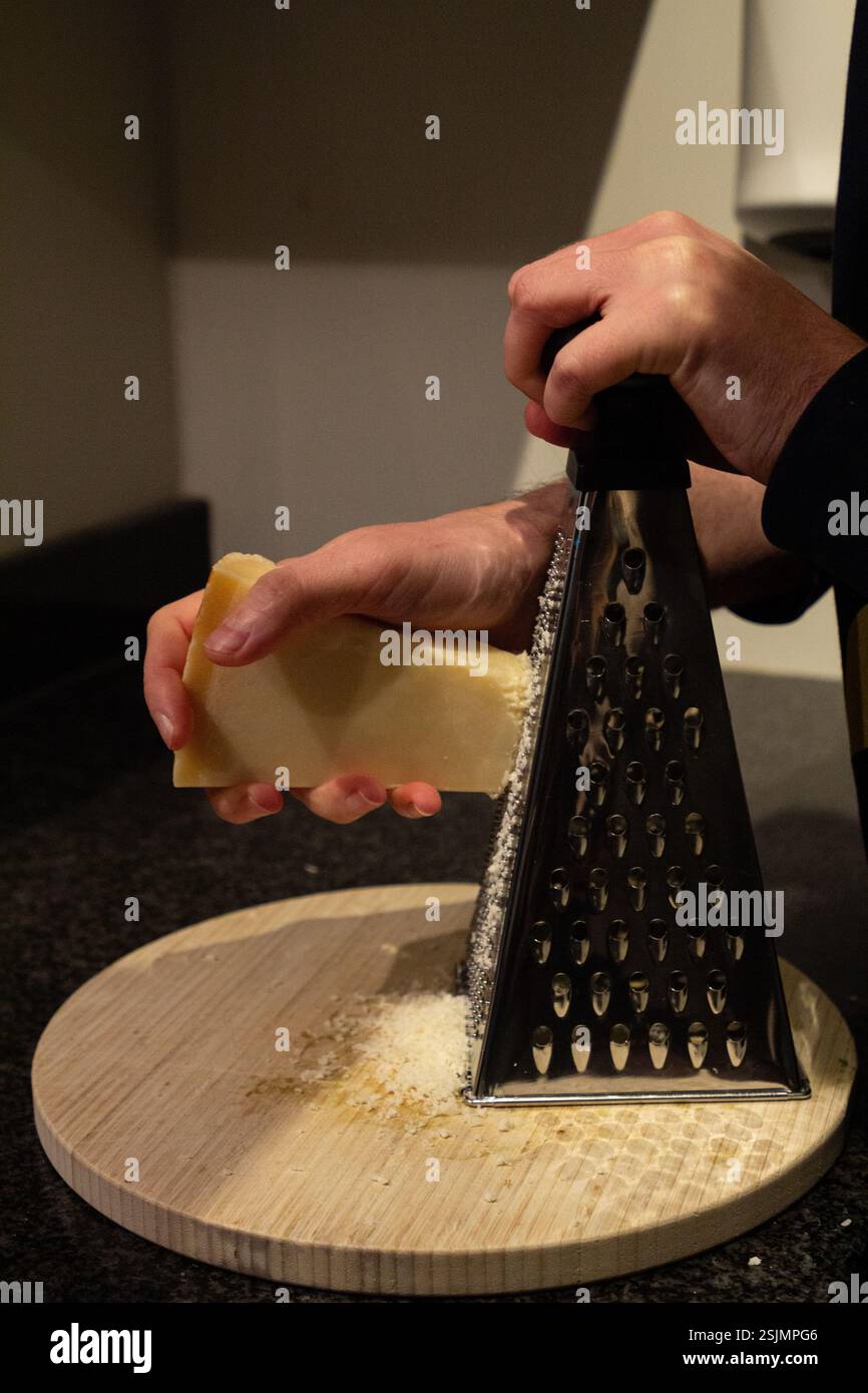 Un primo piano di una mano che utilizza una grattugia per macinare il parmigiano fresco, con il formaggio che cade su una scrivania, mostrando un processo culinario in azione. Foto Stock