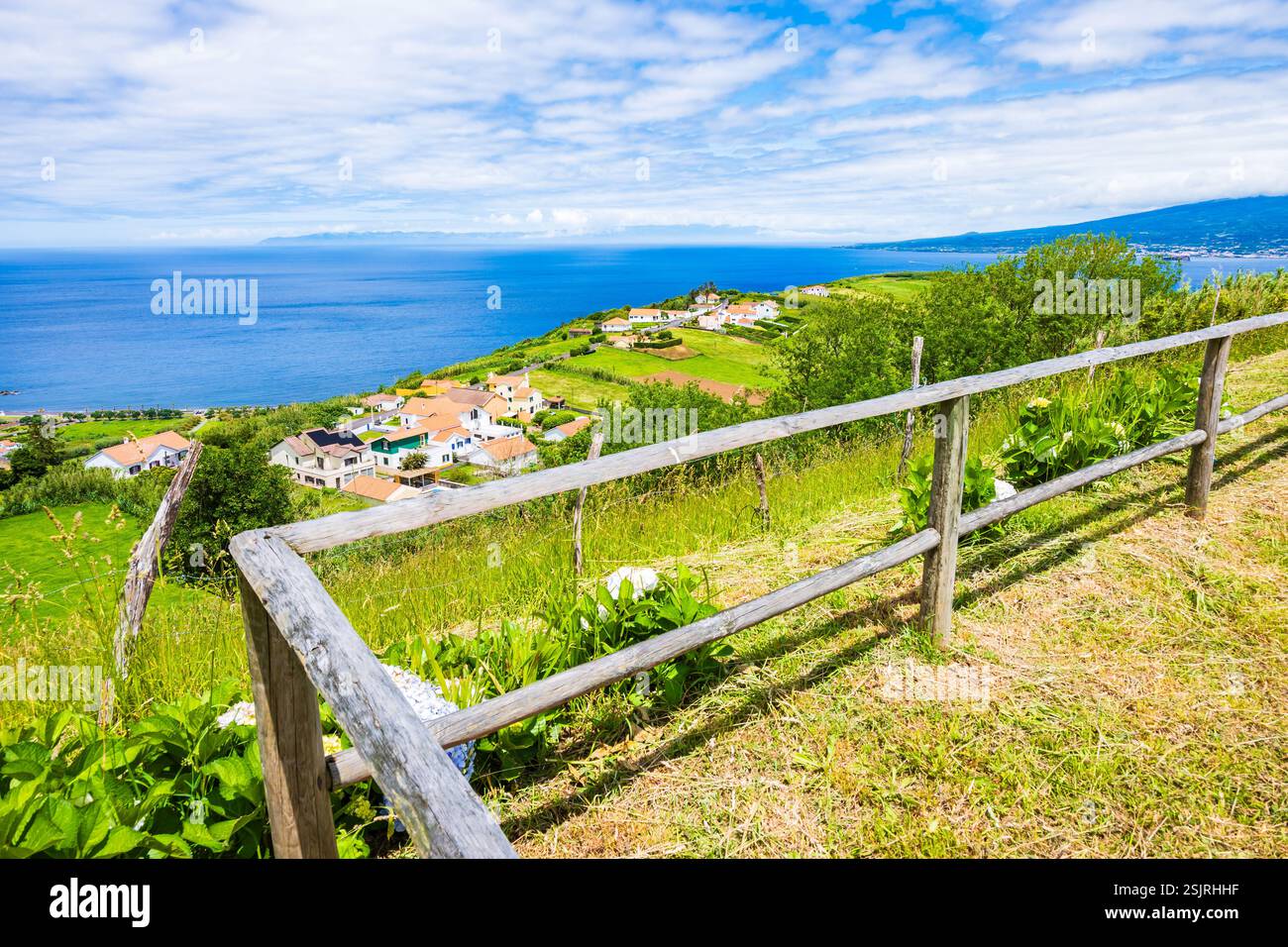 Recinzione in legno su un campo verde in un paesaggio tropicale rurale con costa oceanica e case sullo sfondo, isola di Faial, Azzorre, Portogallo Foto Stock